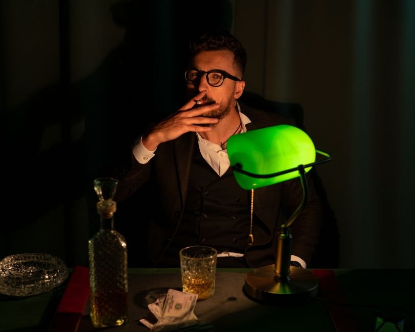 Banker Who Once Owned America — A sophisticated man smoking, seated by a green banker lamp with whiskey and cash, exuding vin
