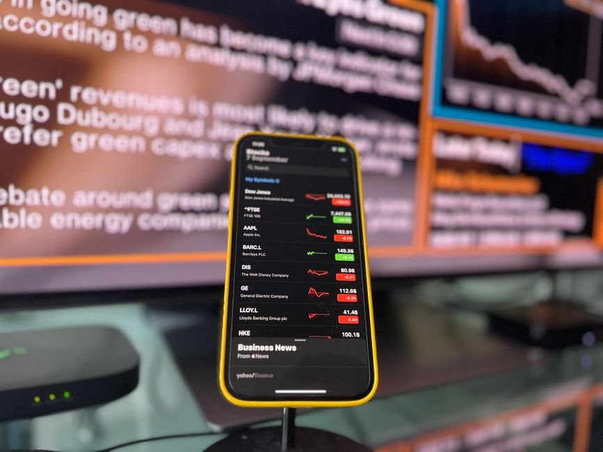 Yahoo — Smartphone showing stock market trends in front of blurred financial news screen indoors.