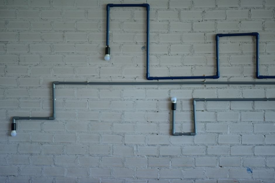 the Phoebus Cartel Engineered Light‑Bulb Death — Industrial style light fixture with pipes and bulbs on a white brick wall. M